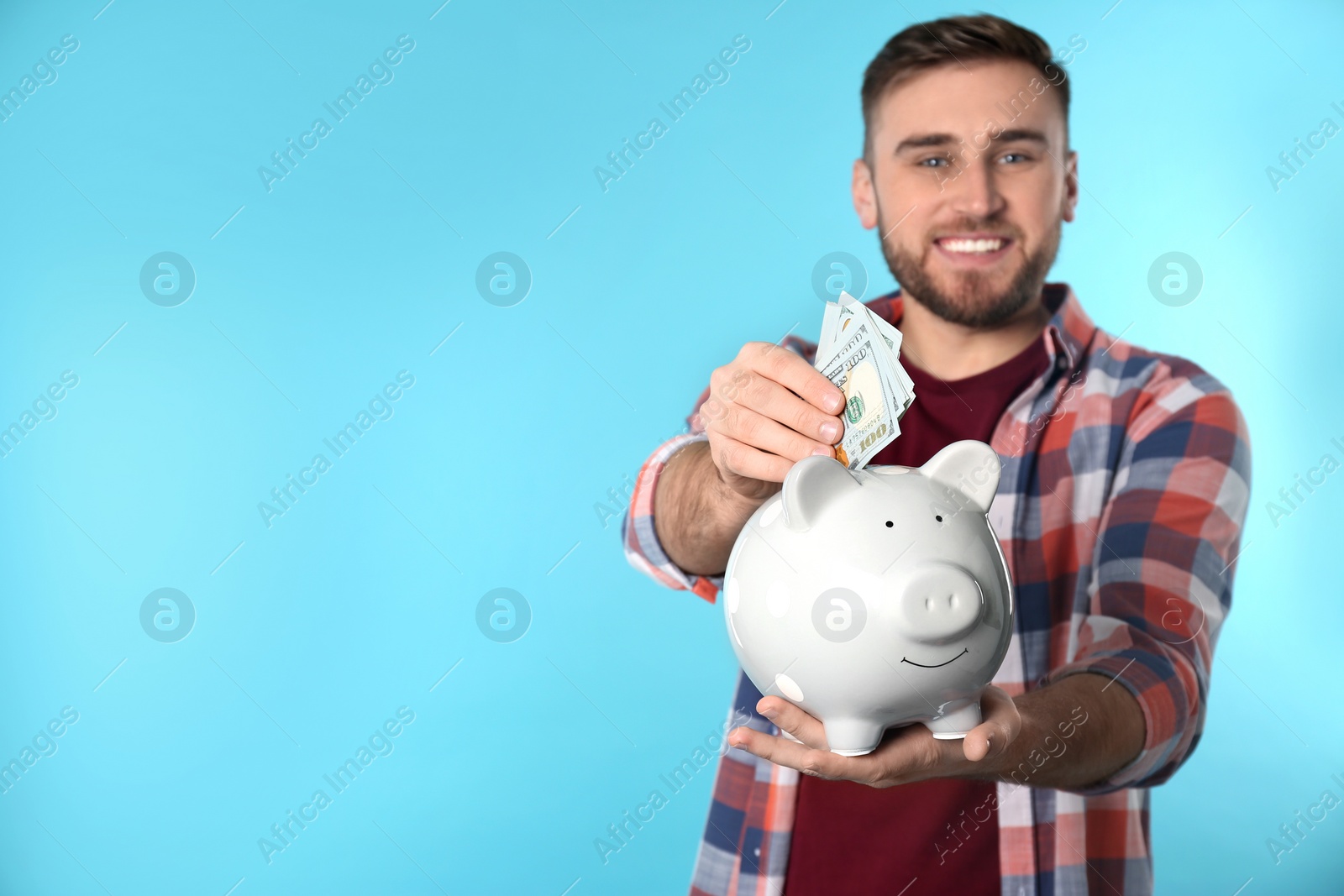 Photo of Happy young man putting money into piggy bank on color background. Space for text