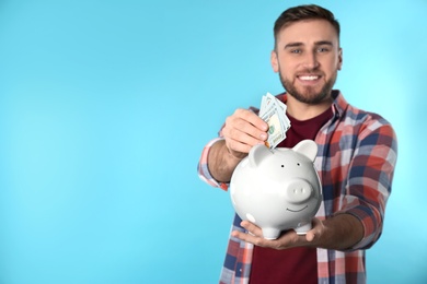 Happy young man putting money into piggy bank on color background. Space for text