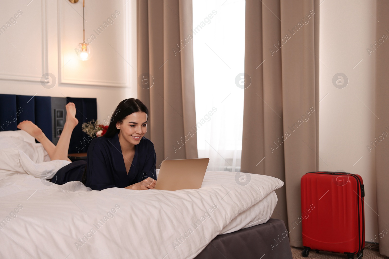 Photo of Beautiful young woman using laptop on bed in hotel room