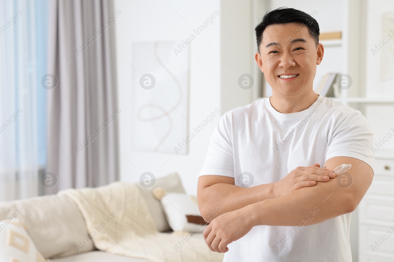 Photo of Handsome man applying body cream onto his arm indoors. Space for text