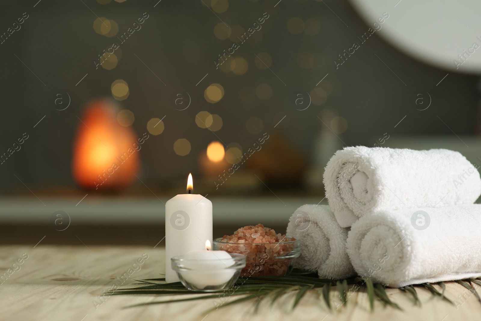 Photo of Spa composition. Rolled towels, sea salt and burning candles on table against blurred lights, space for text