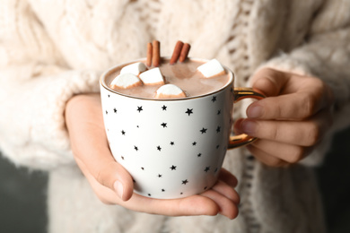 Photo of Woman holding cup of aromatic cocoa with marshmallows and cinnamon sticks, closeup