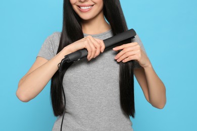 Happy woman using hair iron on light blue background, closeup