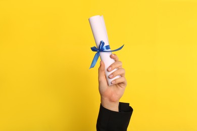Photo of Student holding rolled diploma with blue ribbon on yellow background, closeup