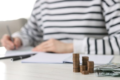 Financial savings. Woman making notes at white wooden table, focus on stacked coins and banknotes