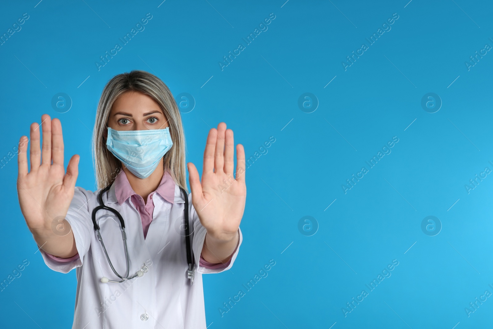 Photo of Doctor in protective mask showing stop gesture on light blue background, space for text. Prevent spreading of coronavirus