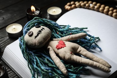 Voodoo doll with pins, book and candles on wooden table, closeup