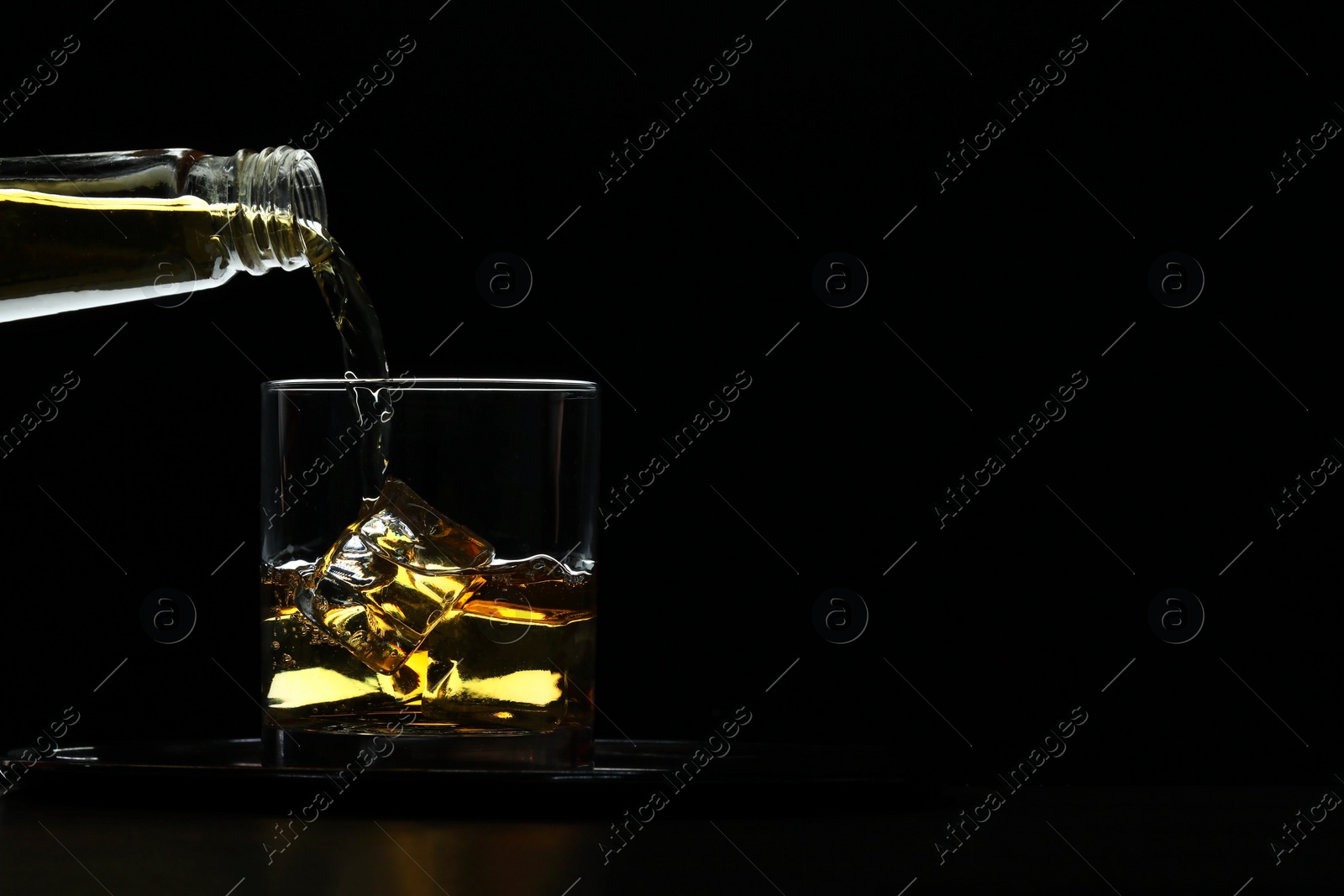 Photo of Pouring whiskey from bottle into glass with ice cubes at table against black background, space for text