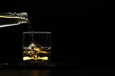 Pouring whiskey from bottle into glass with ice cubes at table against black background, space for text