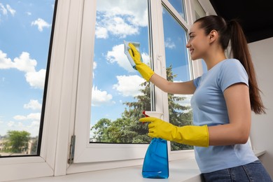 Photo of Happy young woman cleaning window glass with rag and spray indoors