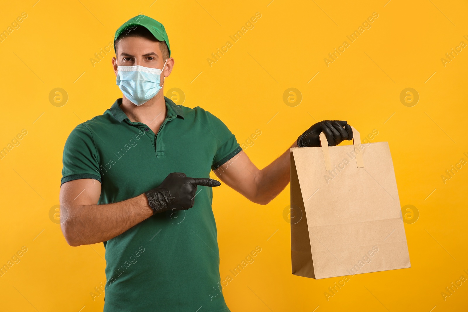 Photo of Courier in medical mask holding paper bag with takeaway food on yellow background. Delivery service during quarantine due to Covid-19 outbreak