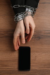Photo of Man with chained hands and smartphone at wooden table, top view. Internet addiction