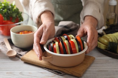 Cooking delicious ratatouille. Woman holding bowl with fresh cut vegetables at white wooden table, closeup