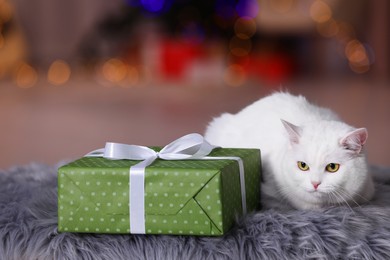 Photo of Christmas atmosphere. Cute cat lying near gift box on fur rug against blurred lights. Space for text