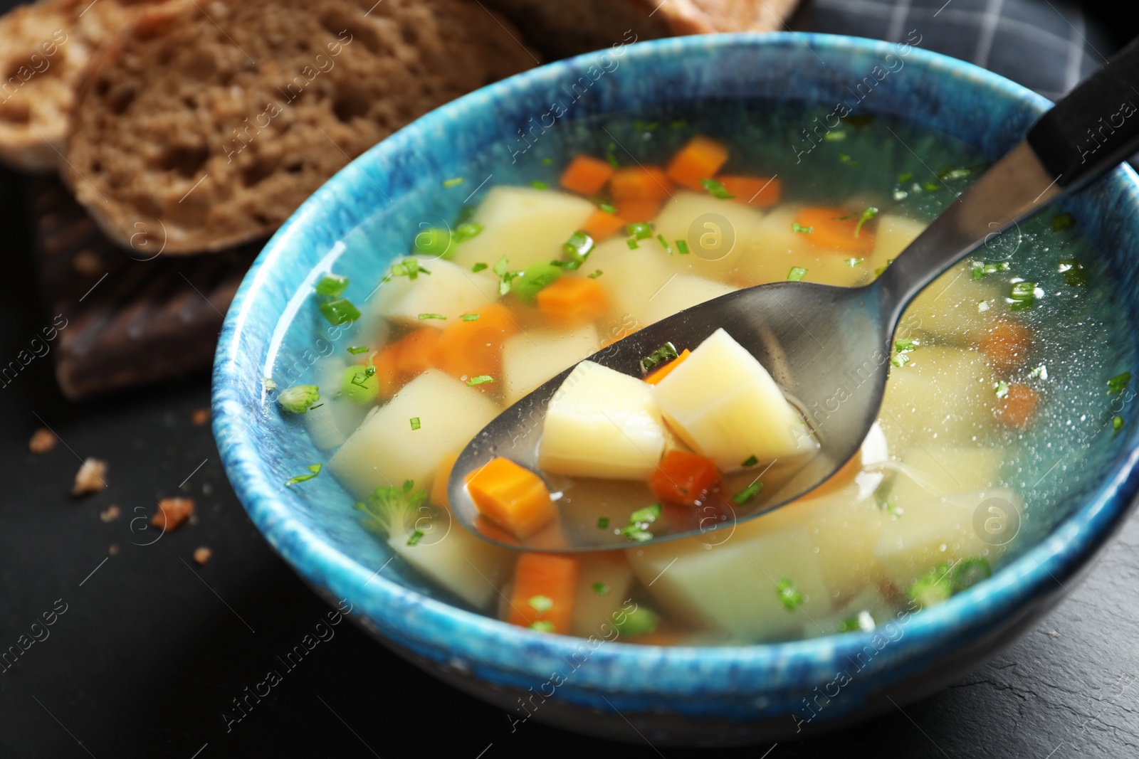 Photo of Spoon of fresh homemade vegetable soup over full bowl on black table, closeup