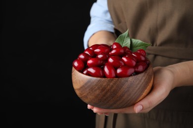 Woman with wooden bowl of fresh ripe dogwood berries on black background, closeup. Space for text