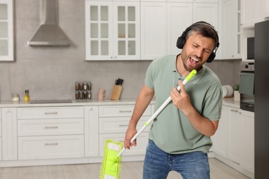 Photo of Happy man in headphones with mop singing while cleaning in kitchen. Space for text