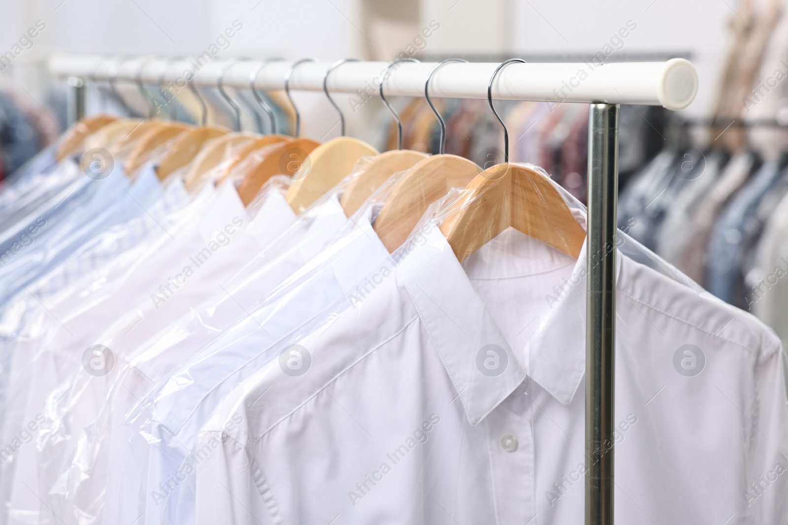 Photo of Dry-cleaning service. Many different clothes in plastic bags hanging on rack indoors, closeup