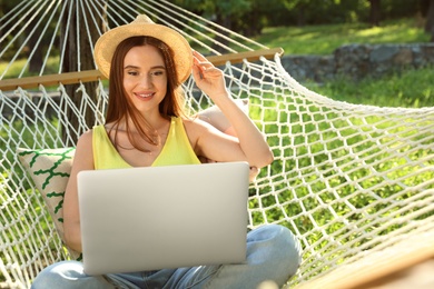Young woman with laptop resting in comfortable hammock at green garden