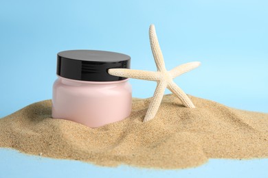 Photo of Jar with cream and starfish on sand against light blue background. Cosmetic product
