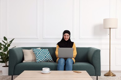 Muslim woman in hijab using laptop on sofa in room