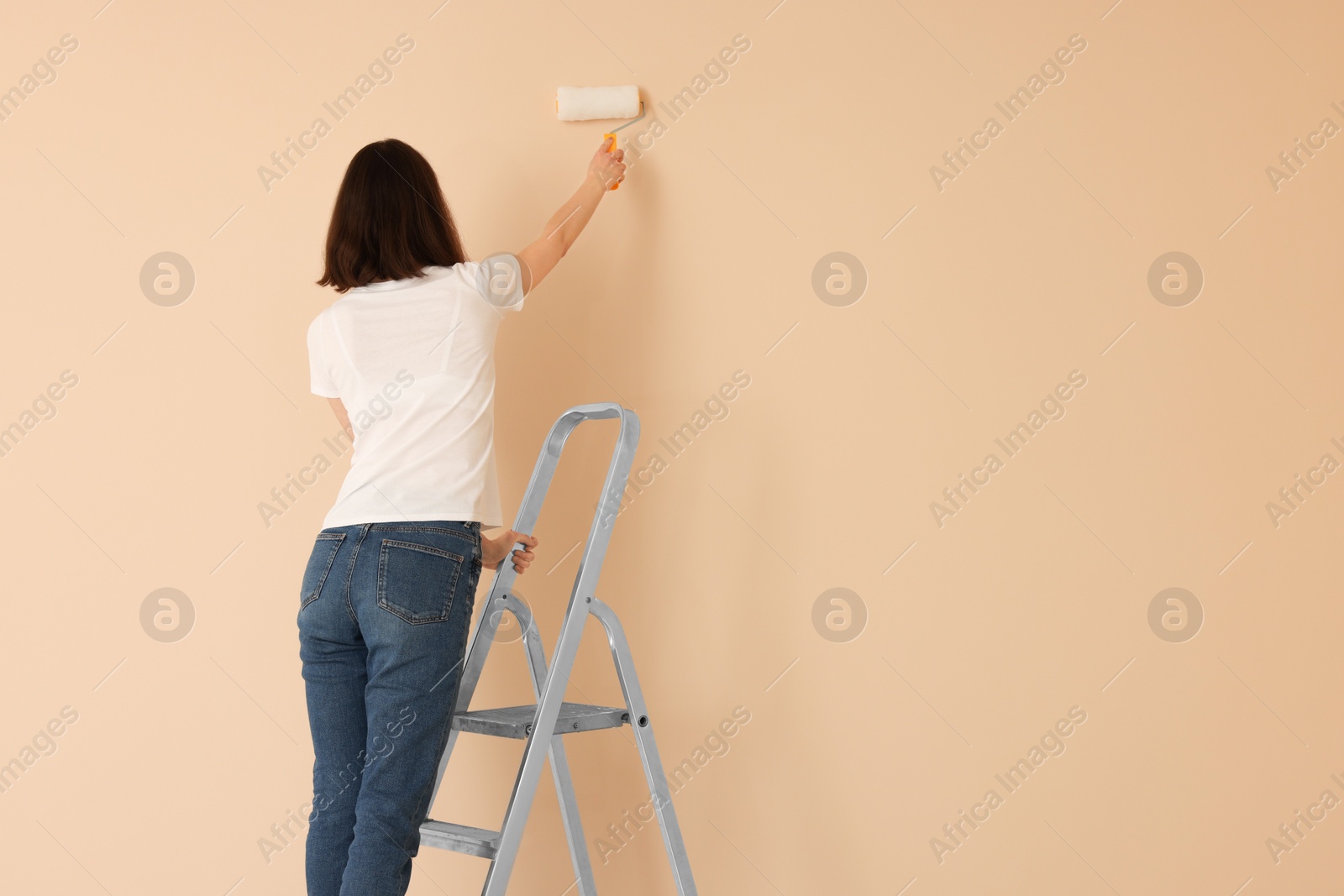 Photo of Young woman painting wall with roller on stepladder indoors, space for text. Room renovation