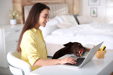 Photo of Young woman working on laptop near her playful dog in home office