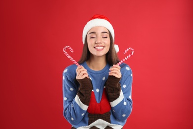 Young woman in Christmas sweater and Santa hat holding candy canes on red background