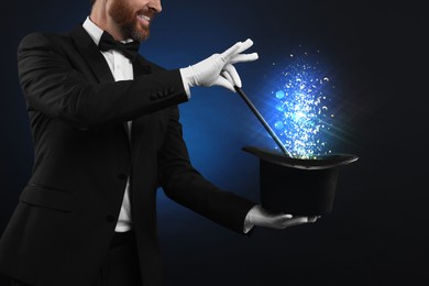 Magician showing trick with wand and top hat on dark background, closeup. Fantastic light coming out of hat