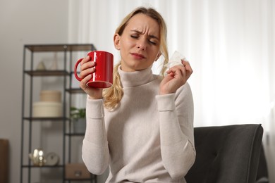 Photo of Sick woman with tissue and cup of drink in armchair at home. Cold symptoms