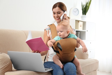 Photo of Woman with her son in baby carrier and book talking on phone at home