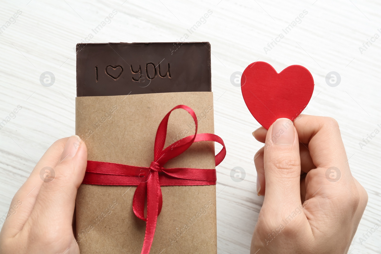 Photo of Woman holding paper heart and chocolate with carved phrase I Love You at white table, closeup