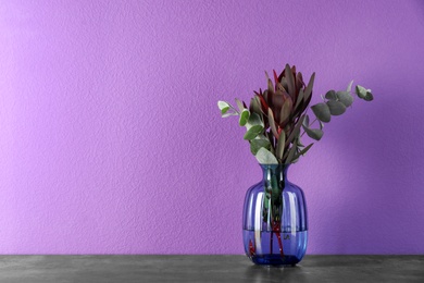 Beautiful bouquet of protea flowers and eucalyptus branches in glass vase on table against purple background