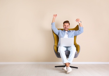 Emotional young man with laptop in armchair near color wall indoors. Space for text