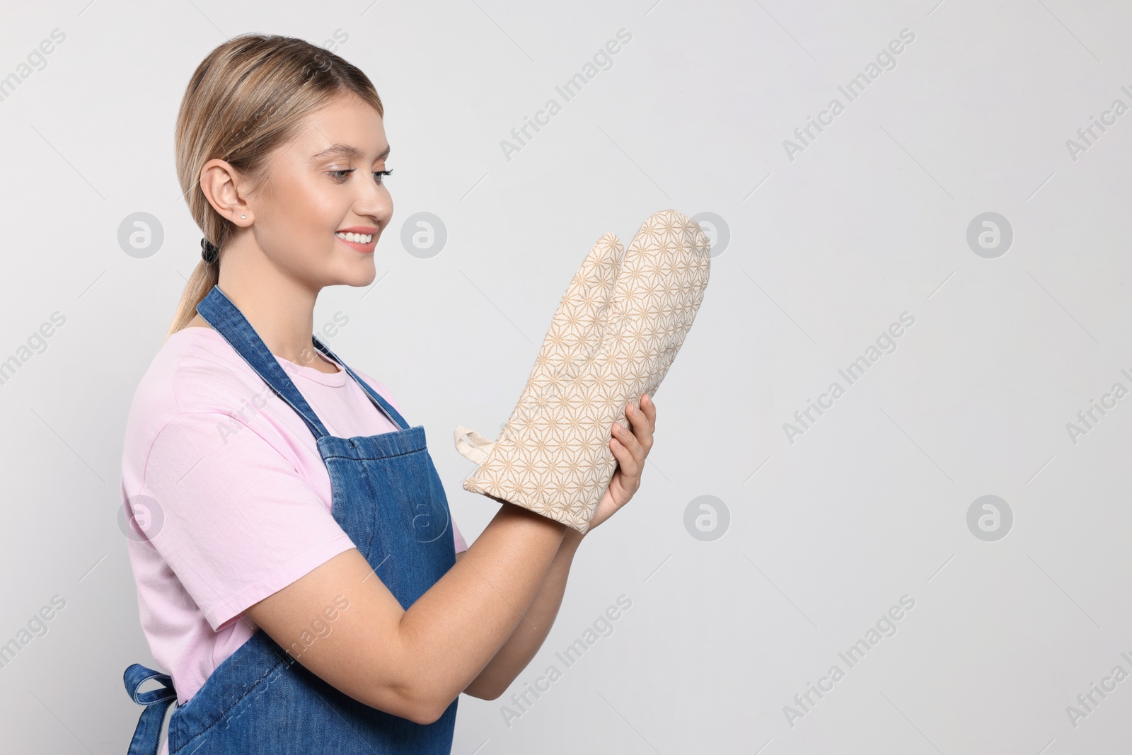 Photo of Beautiful young woman in denim apron and oven glove on light grey background. Space for text