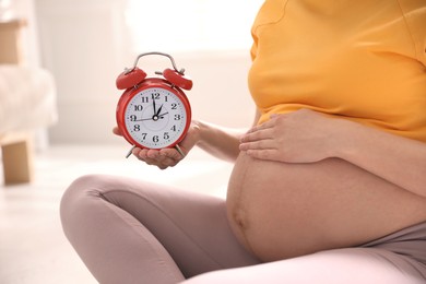 Photo of Young pregnant woman holding alarm clock near her belly at home, closeup. Time to give birth