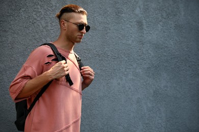 Handsome young man in stylish sunglasses and backpack near grey wall, space for text