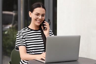 Happy young woman using modern laptop and talking on smartphone at table outdoors