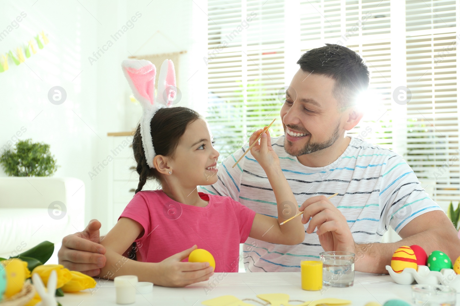 Photo of Happy daughter with bunny ears headband and her father having fun while painting Easter egg at home