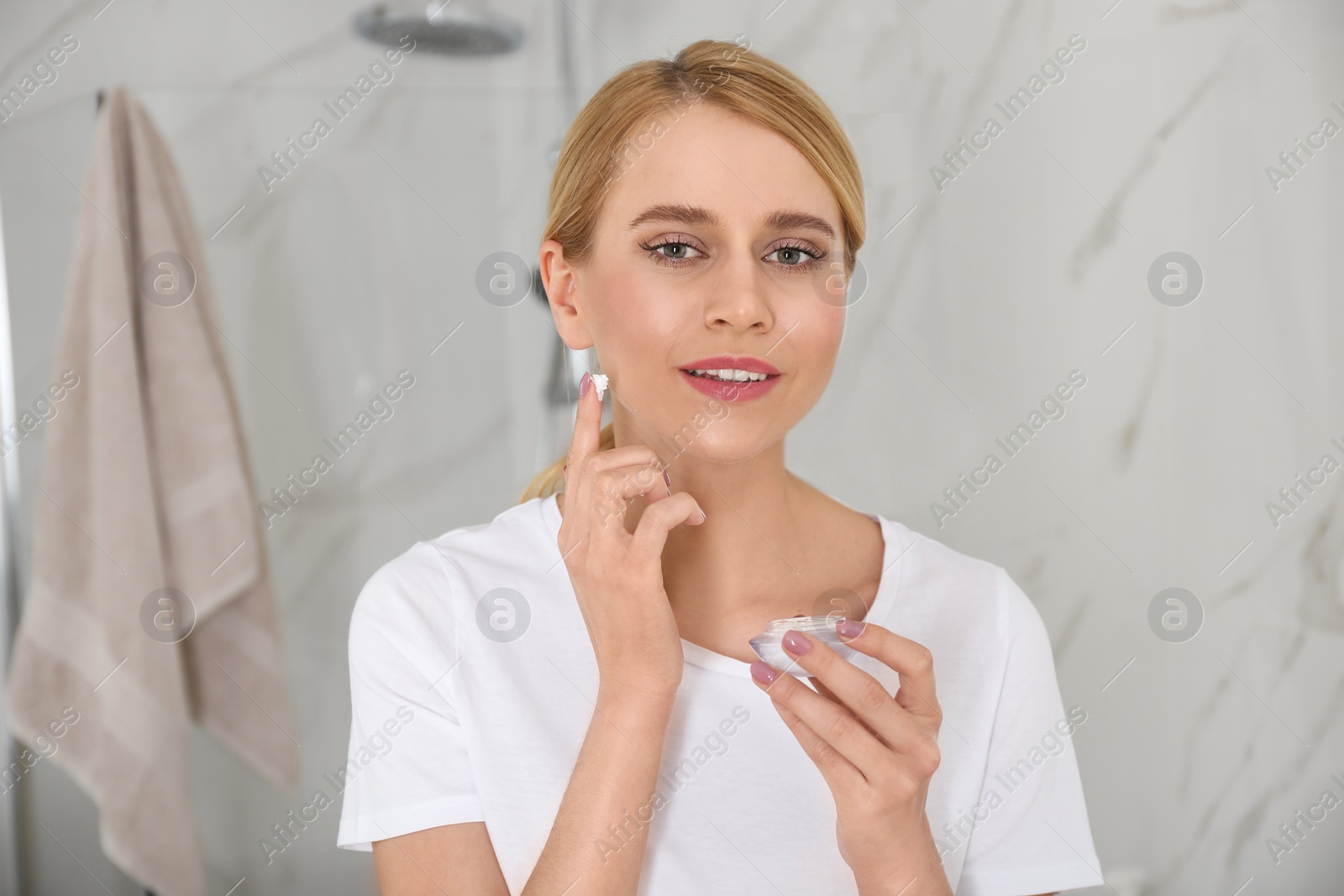 Photo of Happy young woman applying cream onto face in bathroom