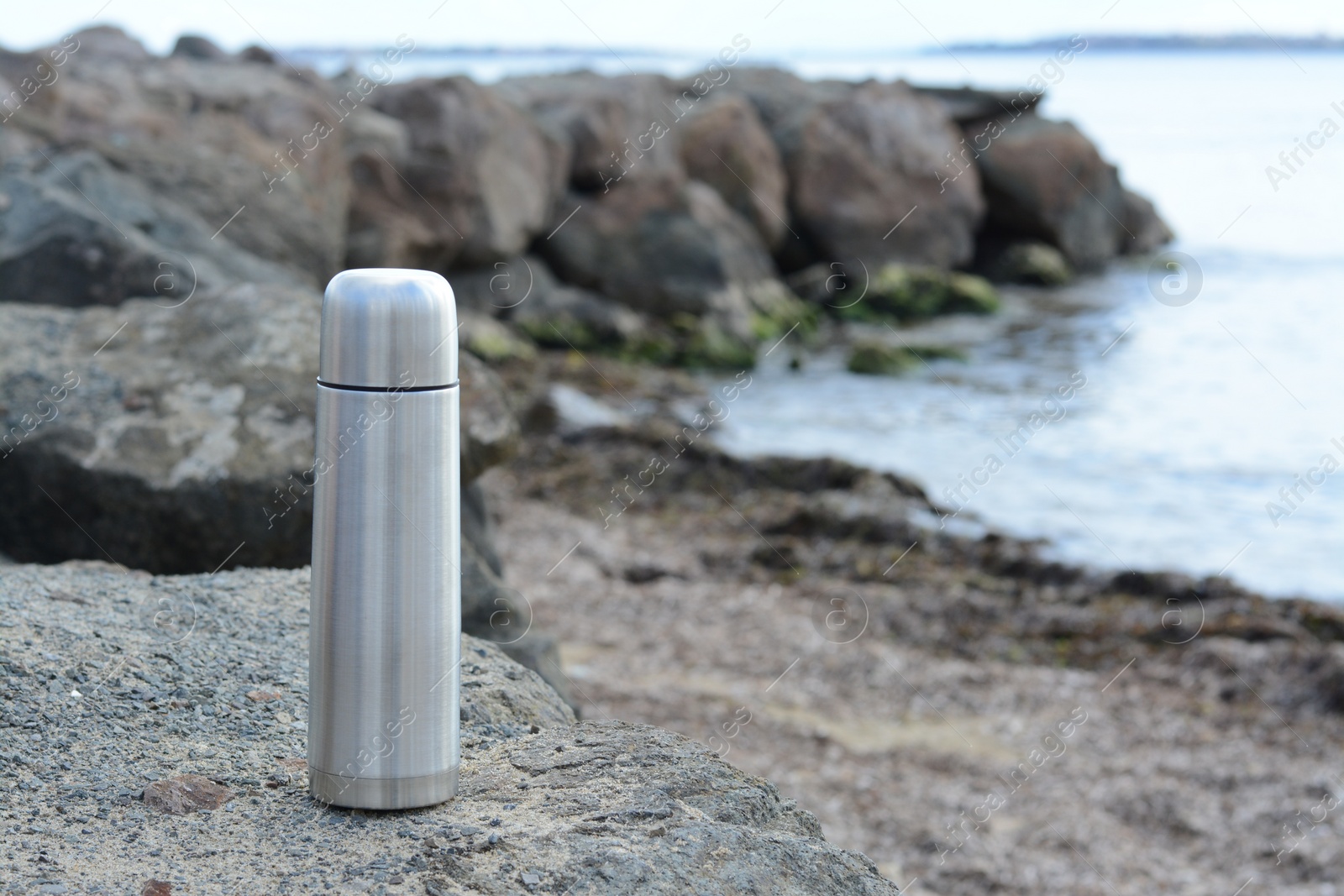 Photo of Metallic thermos with hot drink on stone near sea, space for text