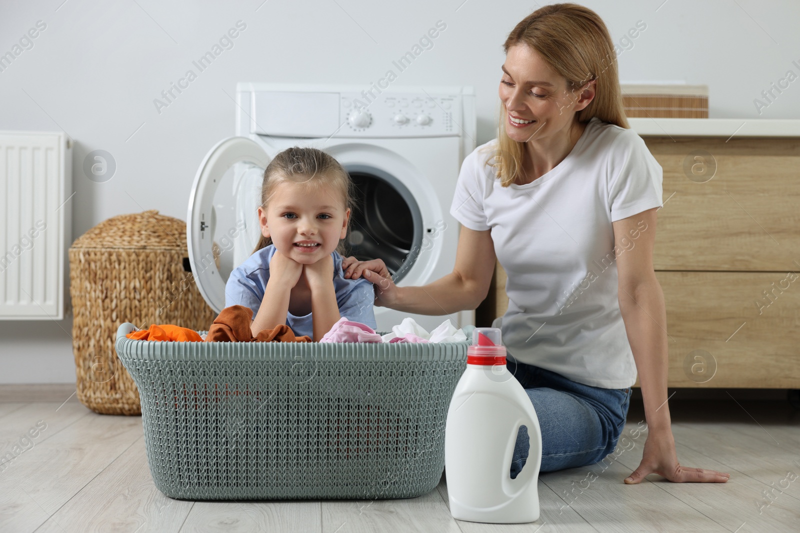 Photo of Mother and daughter sitting on floor near basket with dirty clothes and fabric softener, space for text