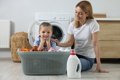 Mother and daughter sitting on floor near basket with dirty clothes and fabric softener, space for text