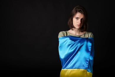 Photo of Sad woman wrapped in Ukrainian flag on black background. Space for text