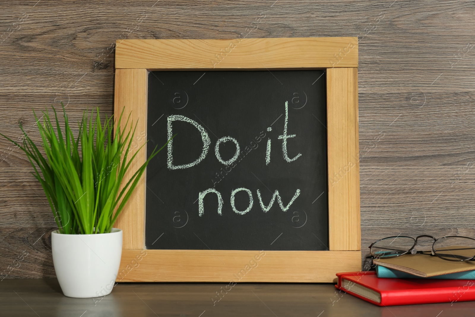 Photo of Small chalkboard with motivational quote Do It Now, stationery and plant on wooden table