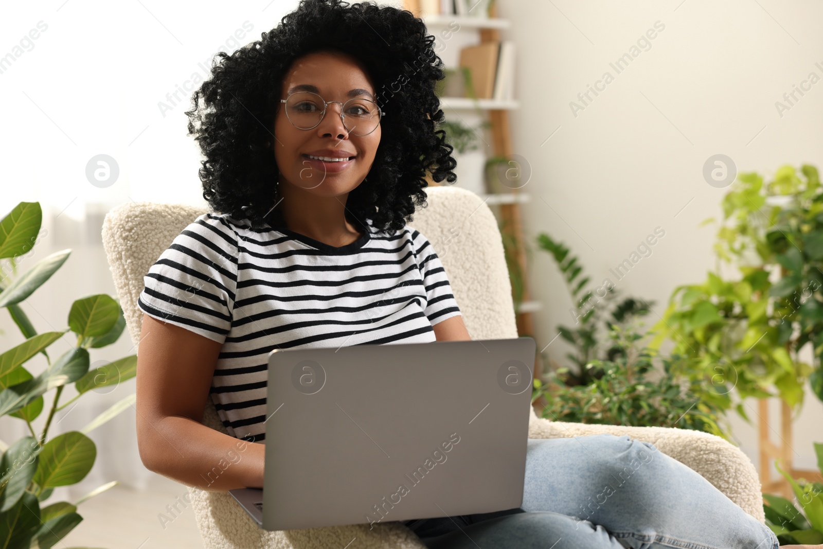 Photo of Relaxing atmosphere. Happy woman with laptop sitting on armchair near houseplants at home