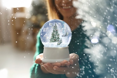 Image of Woman holding snow globe with Christmas tree at home, focus on hands