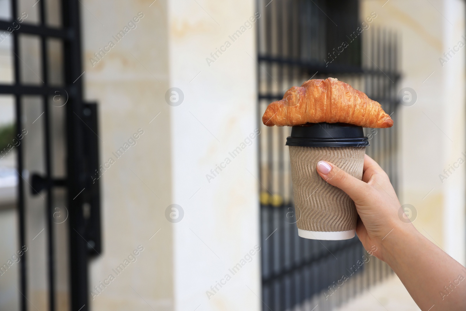 Photo of Woman holding croissant and paper cup in hand outdoors, closeup. Space for text