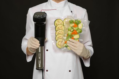 Chef holding sous vide cooker and vegetables in vacuum packs on black background, closeup