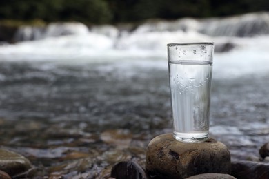 Glass of fresh water on stone near river, space for text